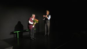 Auf der Bühne: Links steht eine Frau mit einem Blumenstrauß. Rechts von ihr ein Mann der applaudiert.